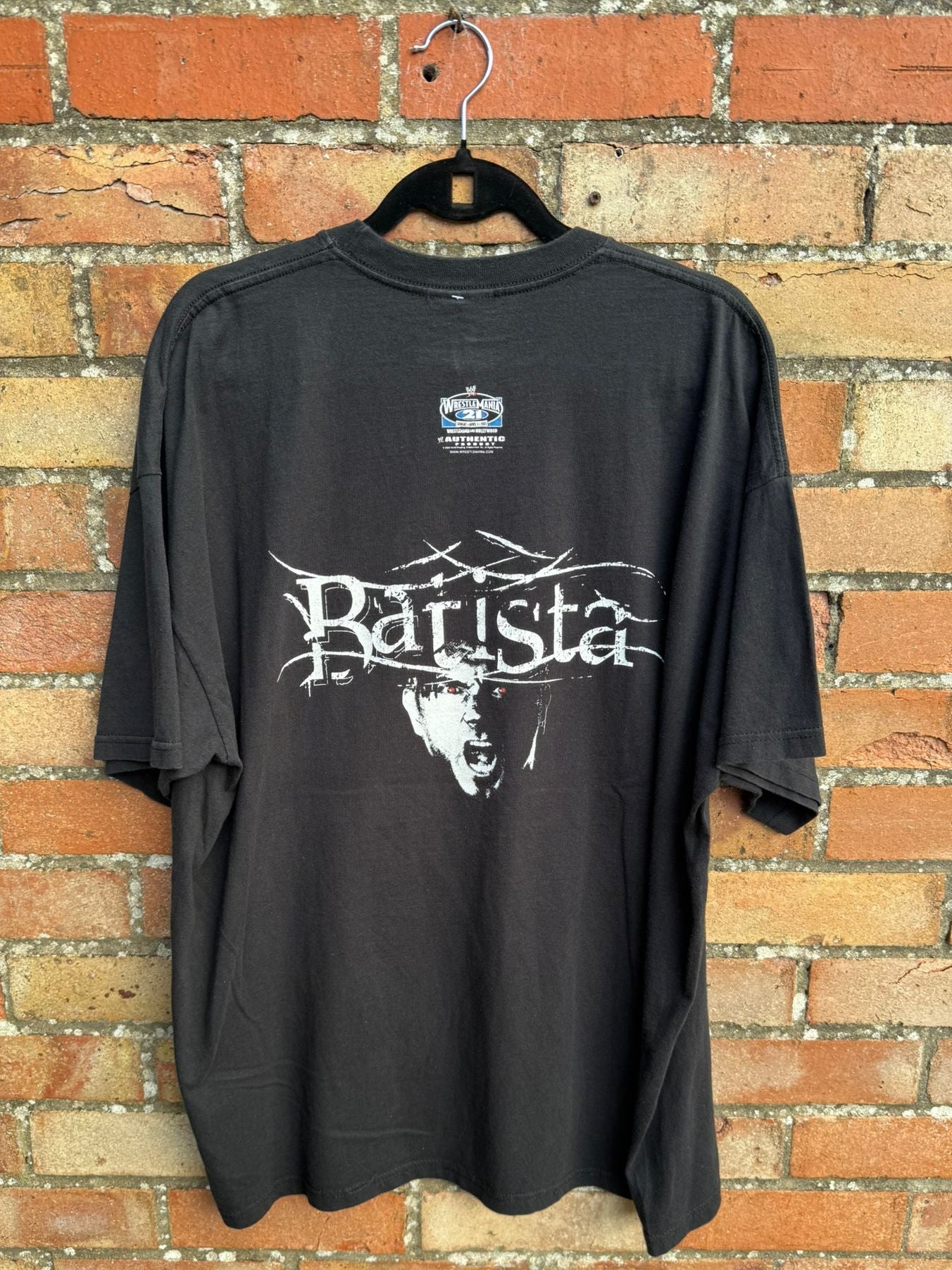 Back view of black t-shirt with 'Batista' design and Wrestlemania 21 logo, on a hanger against a brick wall.