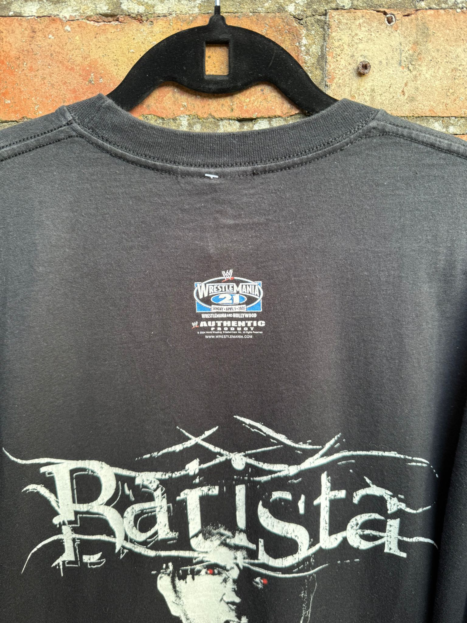 Back of black t-shirt with 'Batista' text and  Wrestlemania 21 logo showing, on a hanger against a brick wall.