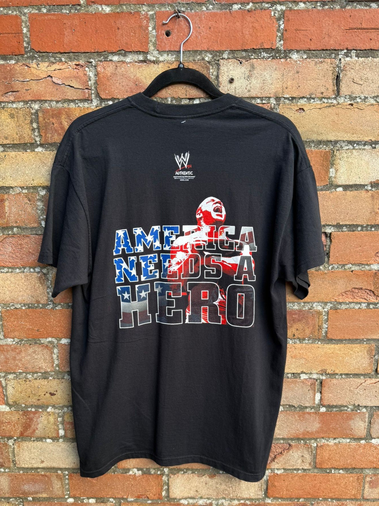 Back view of black t-shirt with 'America Needs a Hero' graphic on a hanger against a brick wall.
