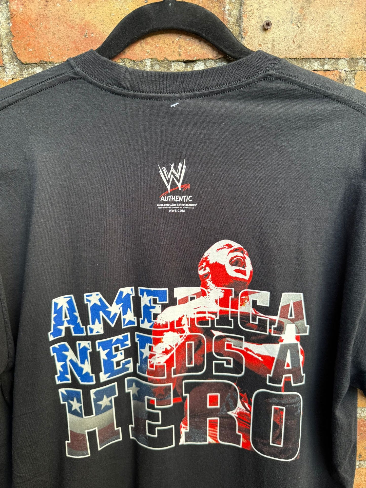 Black t-shirt with 'America Needs a Hero' graphic and WWE logo on a hanger against a brick wall.