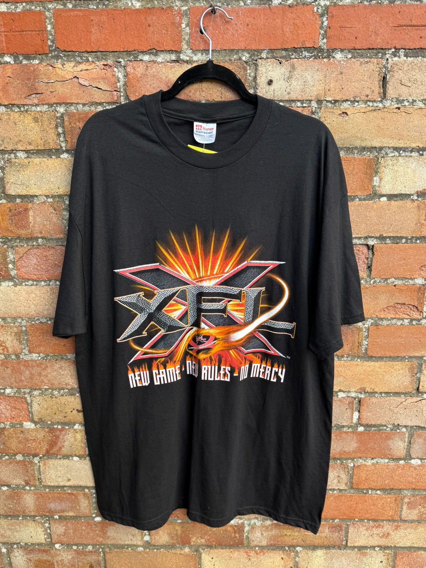 Black t-shirt with large XFL print and text New Game New Rules No Mercy, hanging against a brick wall