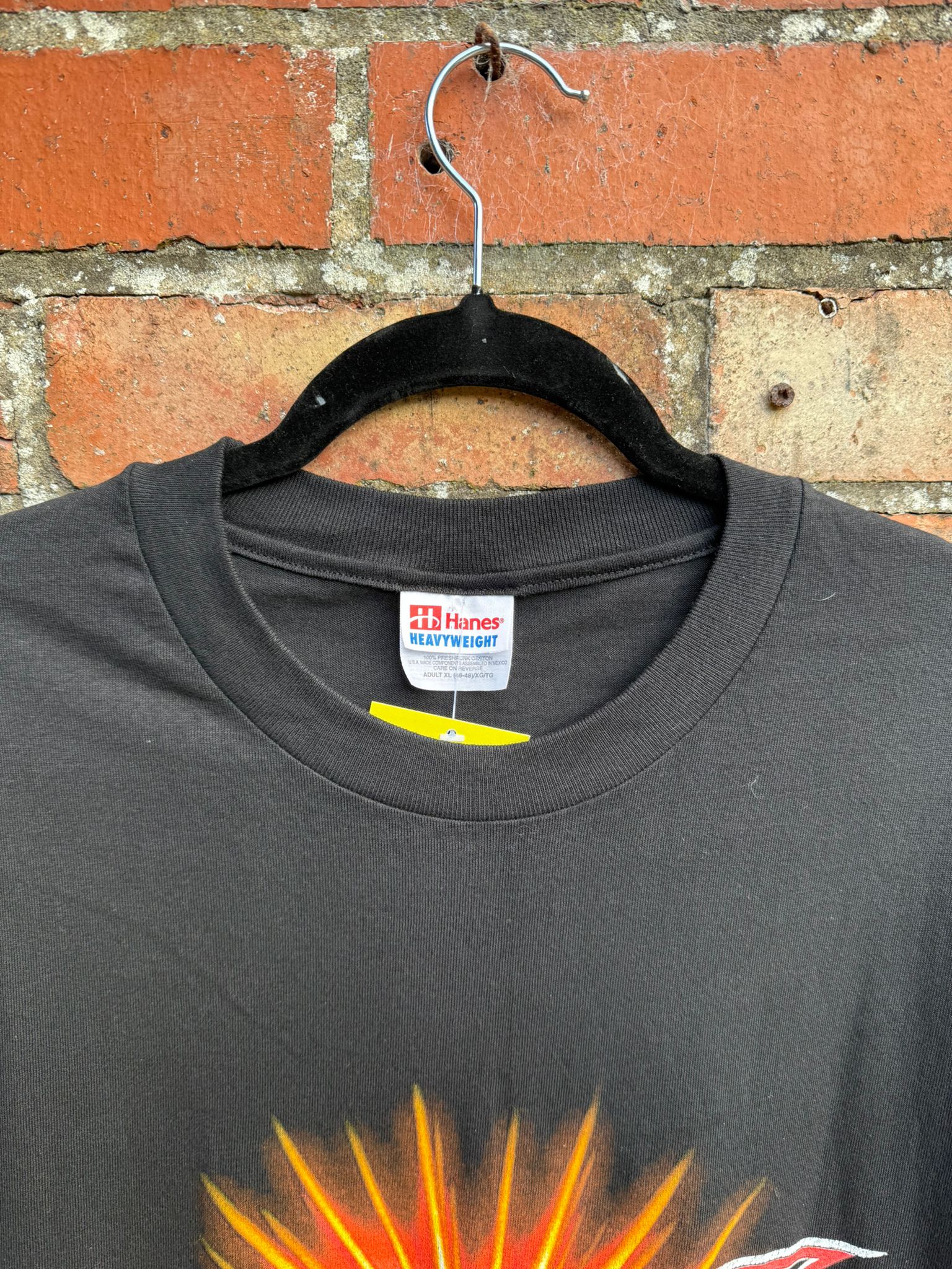Black t-shirt showing a Hanes Heavyweight tag and with a colorful design on a hanger against a brick wall.