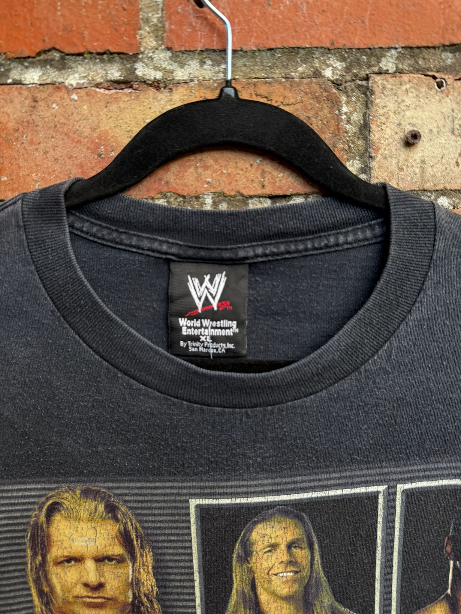 Black t-shirt with WWE logo and images of wrestlers on a hanger against a brick wall.