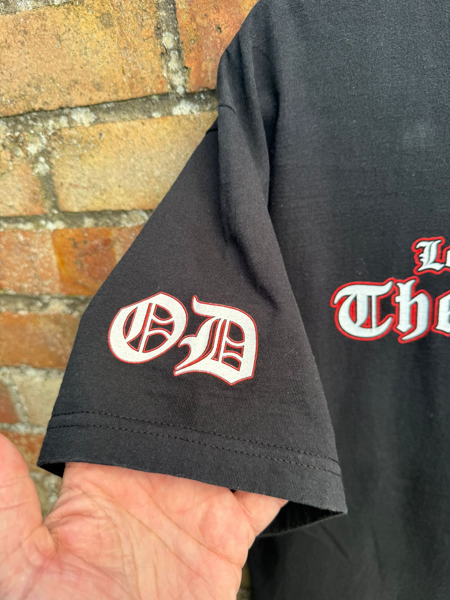 Black t-shirt with red and white text held against a brick wall.