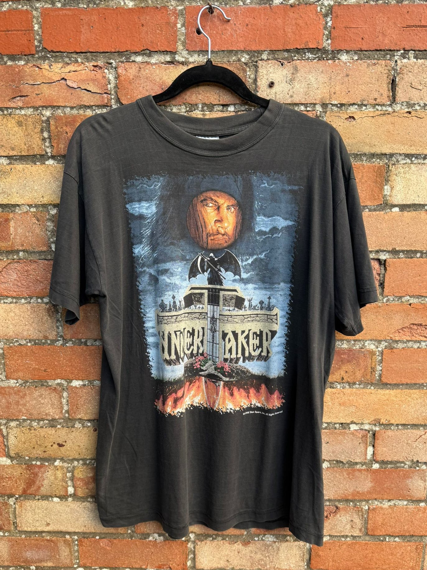 Black t-shirt with The Undertaker graphic design on a hanger against a brick wall