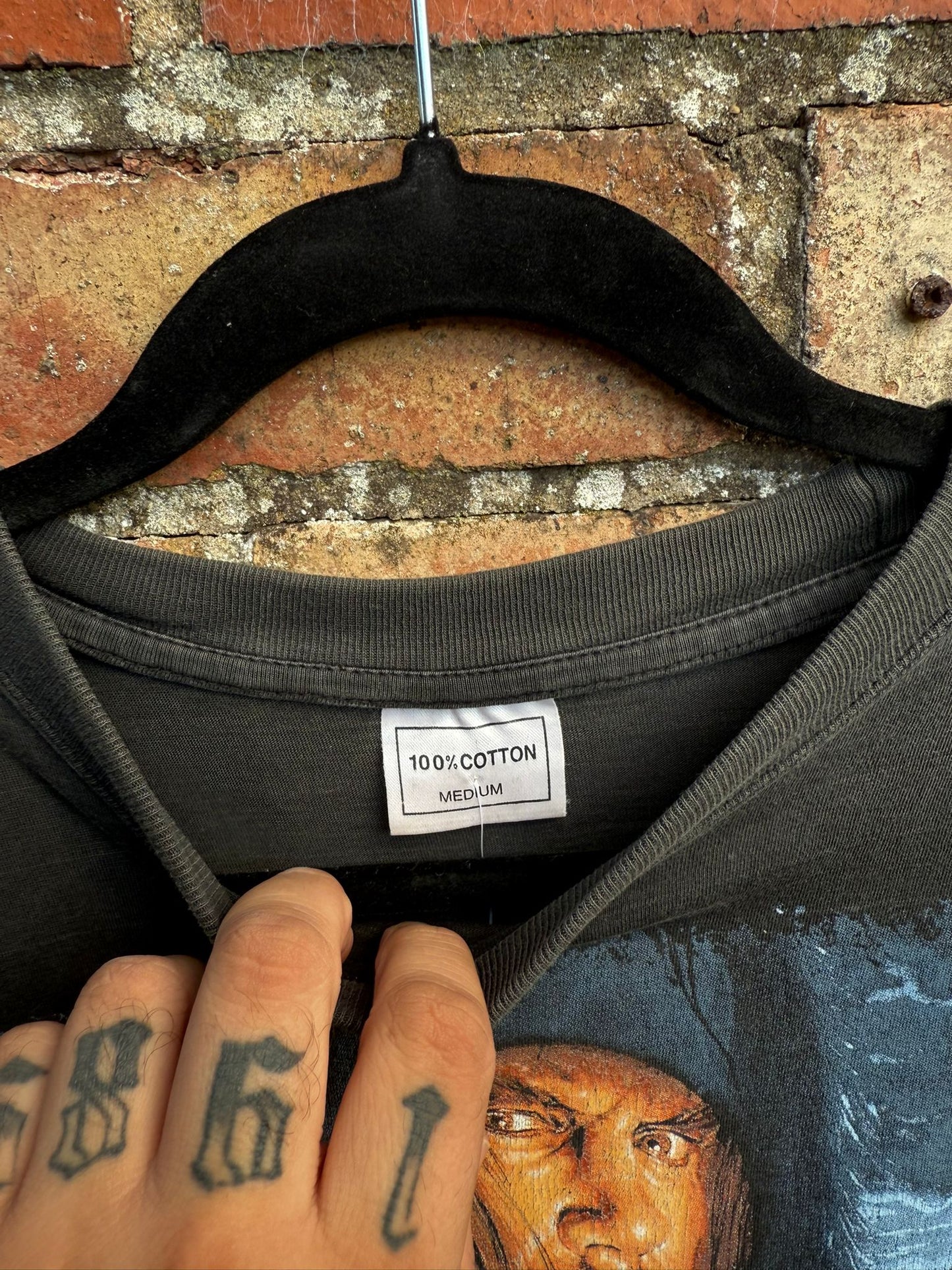 Black t-shirt with a 100% cotton and Medium tag and graphic design on a hanger against a brick wall.