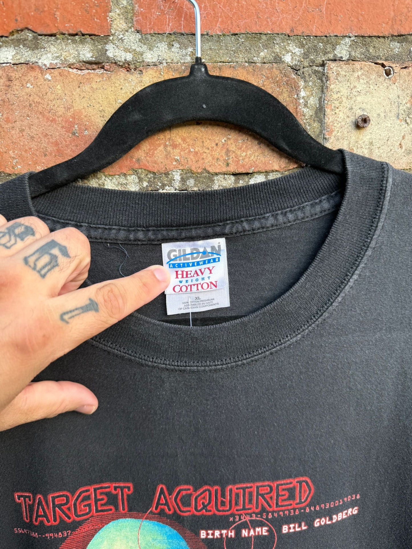 close up of Gildan tag on a black t-shirt with 'Target Acquired' text on a hanger against a brick wall.