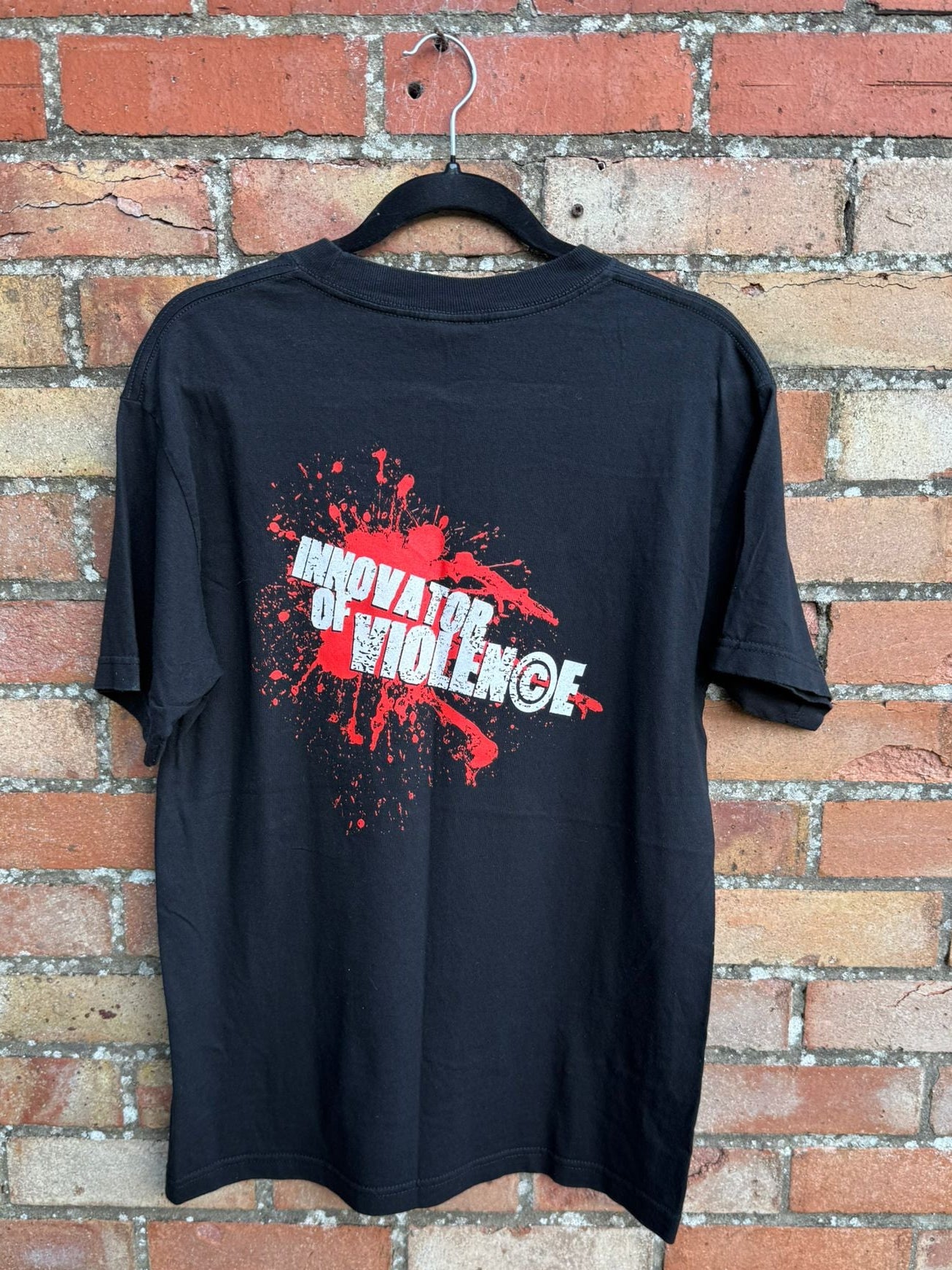 Back of a black t-shirt with red and white graphic design with Innovator of Violence text, on a hanger against a brick wall.