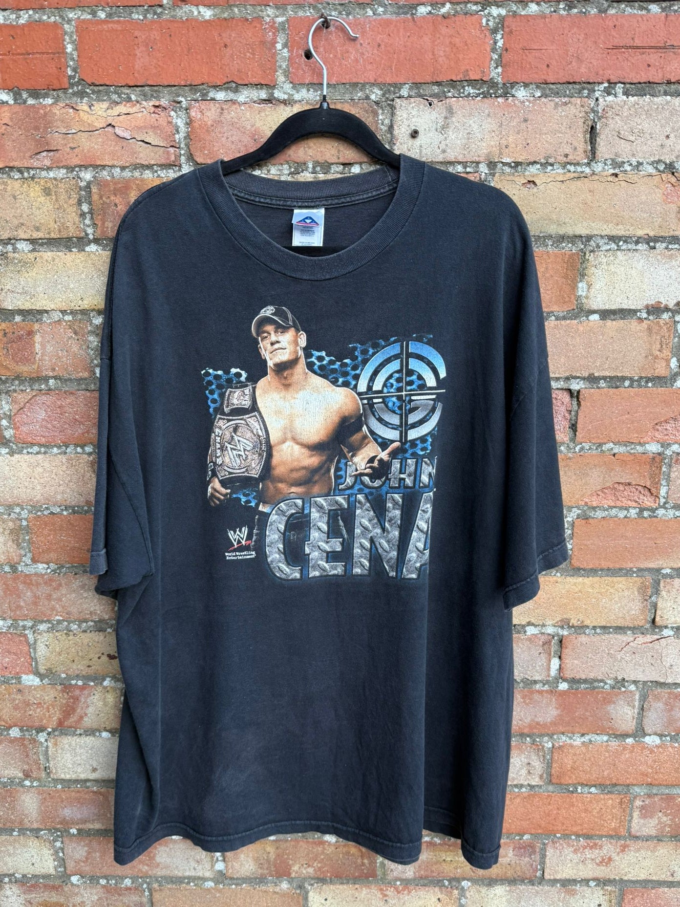 Black t-shirt with a graphic of wrestler John Cena holding a championship belt, hanging against a brick wall.