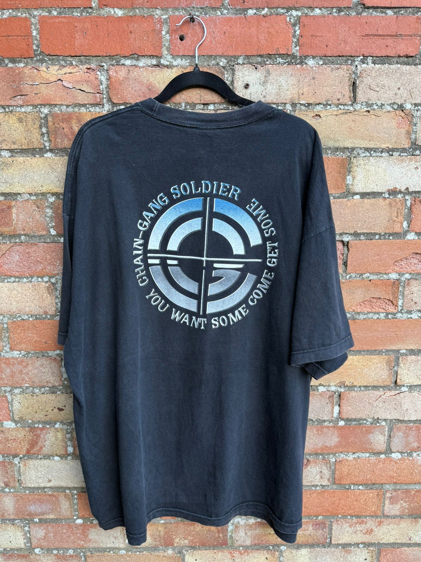 Black t-shirt with graphic design and text chain-gang soldier on a hanger against a brick wall