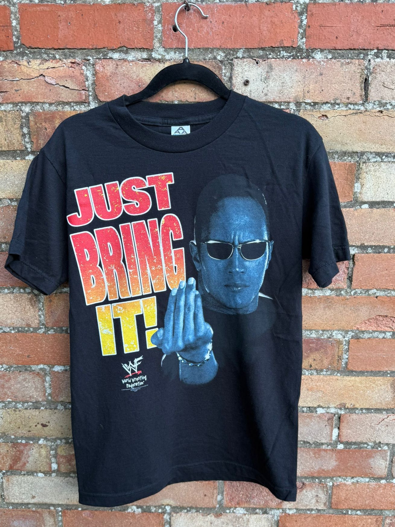 Black t-shirt with image of The Rock and text Just Bring It, hangin against a brick wall