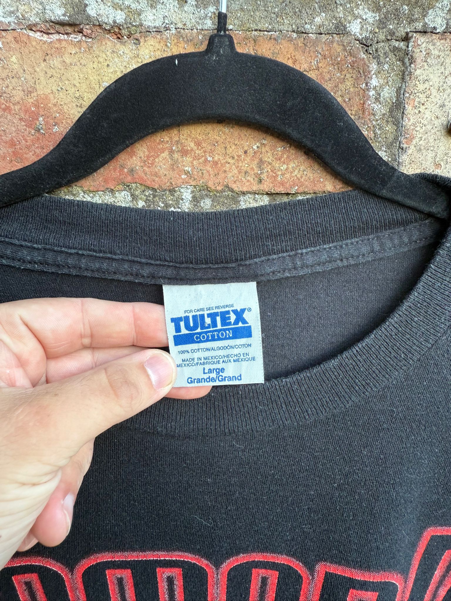 Black t-shirt with a Tultex label held by a hand against a brick wall background