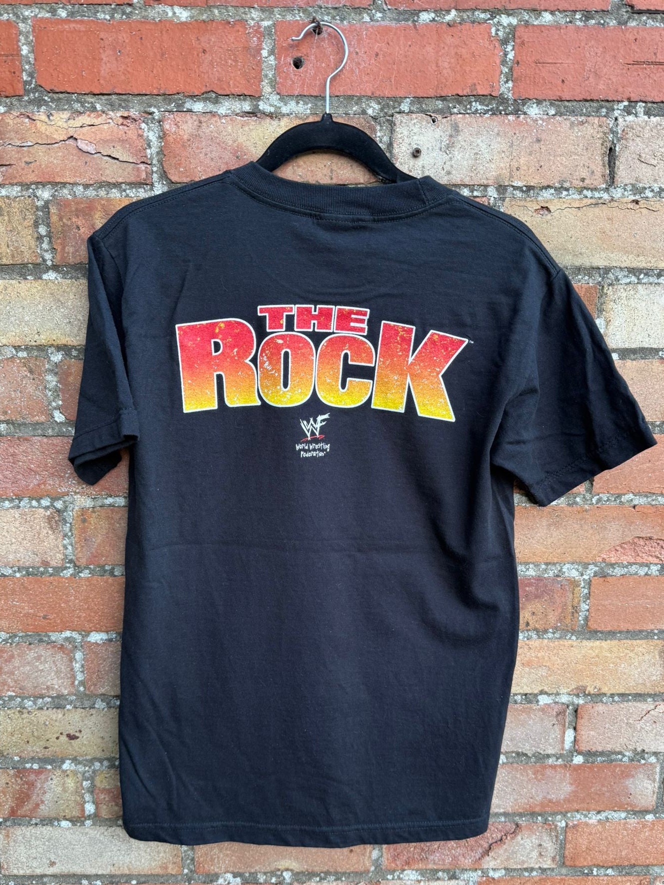 Black t-shirt with 'The Rock' graphic on a brick wall background