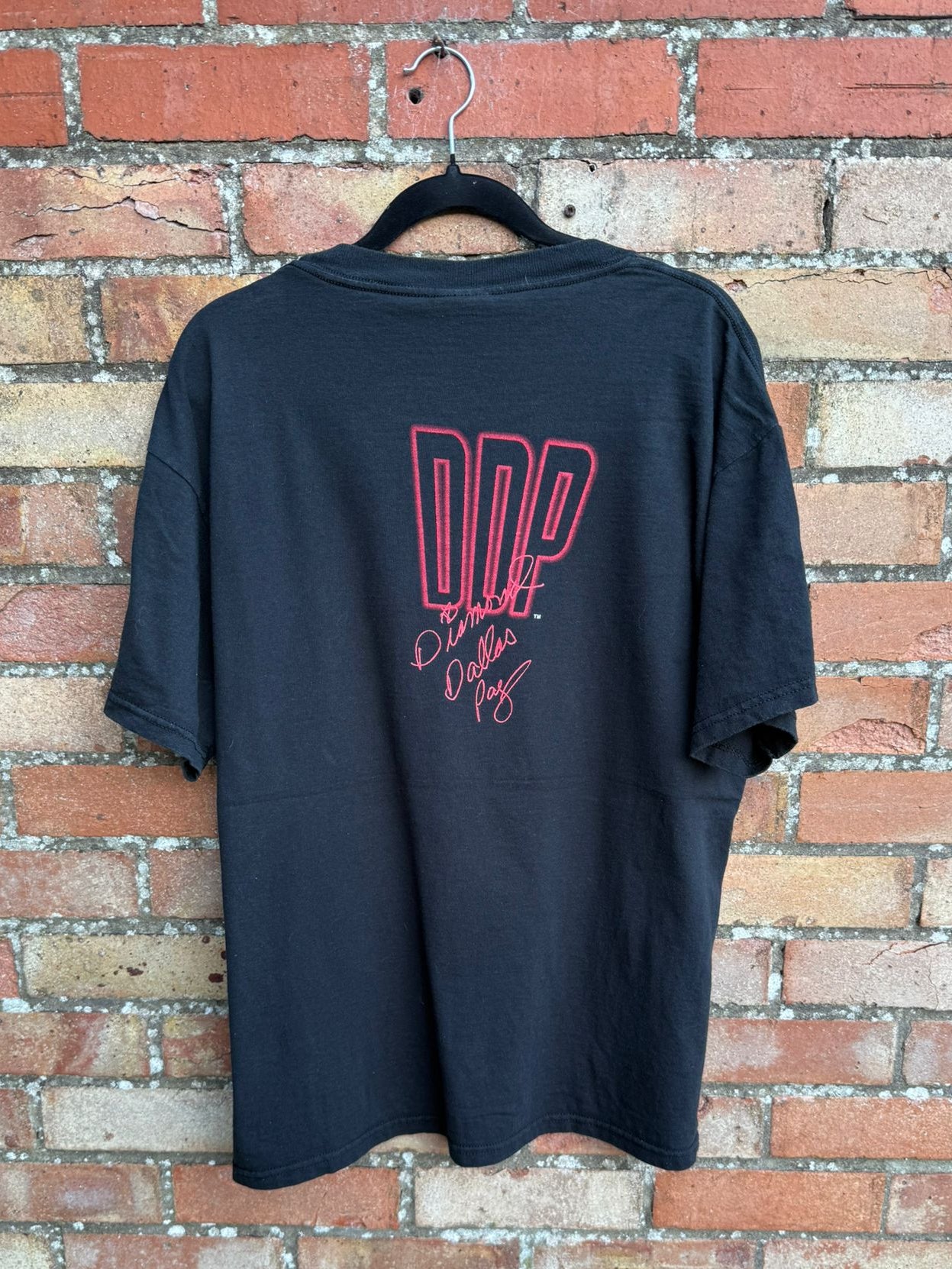 Black t-shirt with red print DDP and signature on a hanger against a brick wall