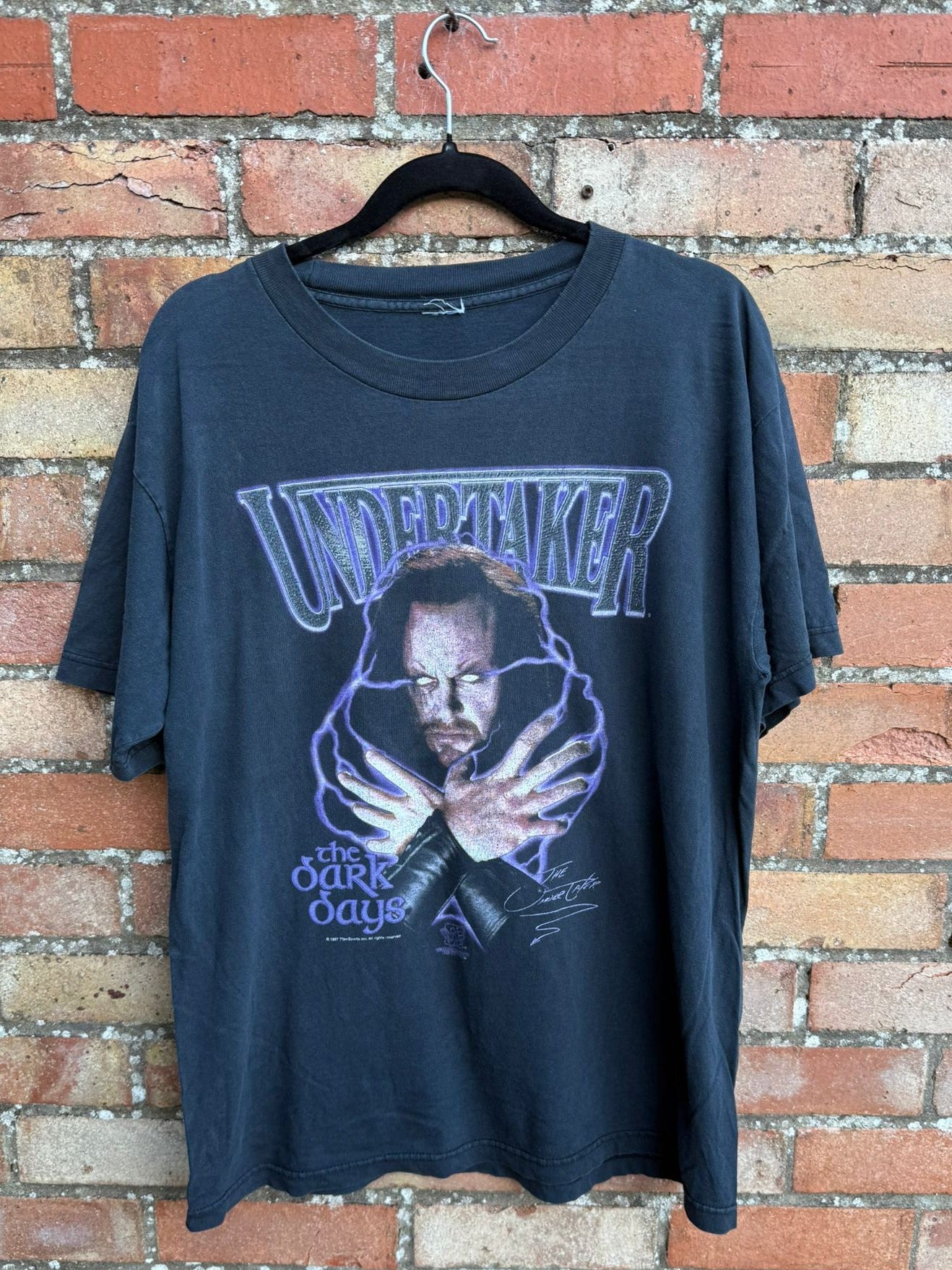 Black t-shirt with graphic design of The Undertaker The Dark Days, on a hanger against a brick wall.