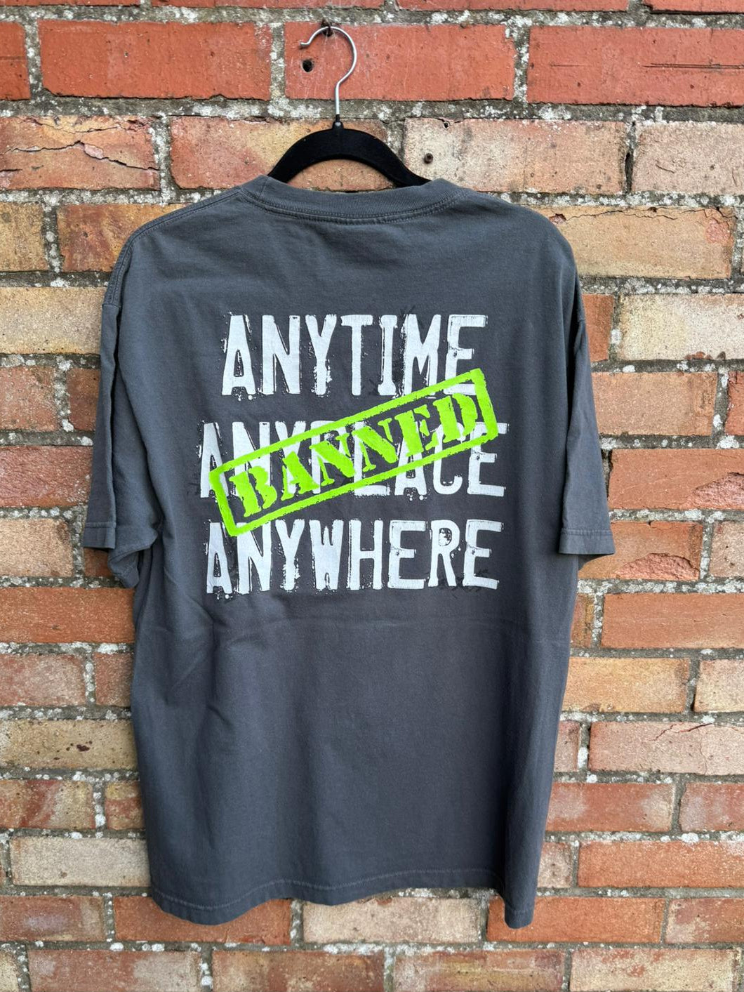 Back of a gray t-shirt with text Anytime Anyplace Anywhere on a hanger against a brick wall