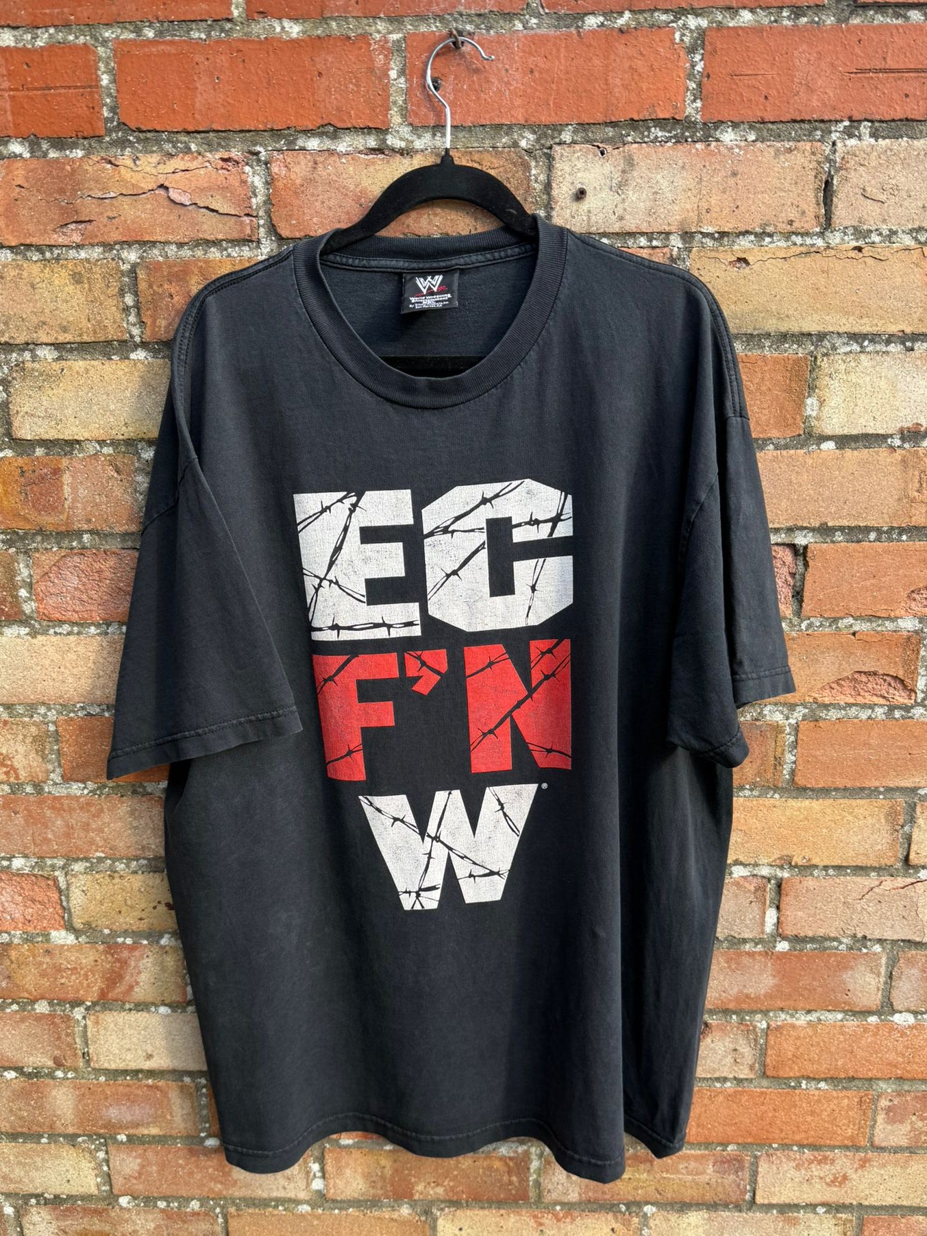 Black t-shirt with graphic text EC F'N W on a hanger against a brick wall