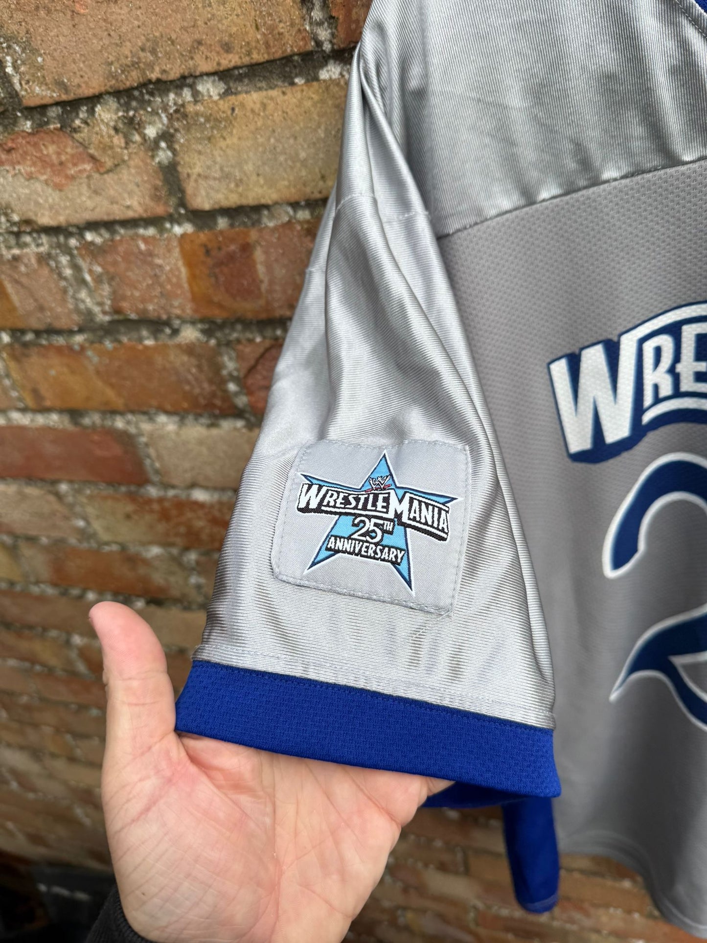 Gray sports jersey with WrestleMania 25 anniversary patch held against a brick wall.