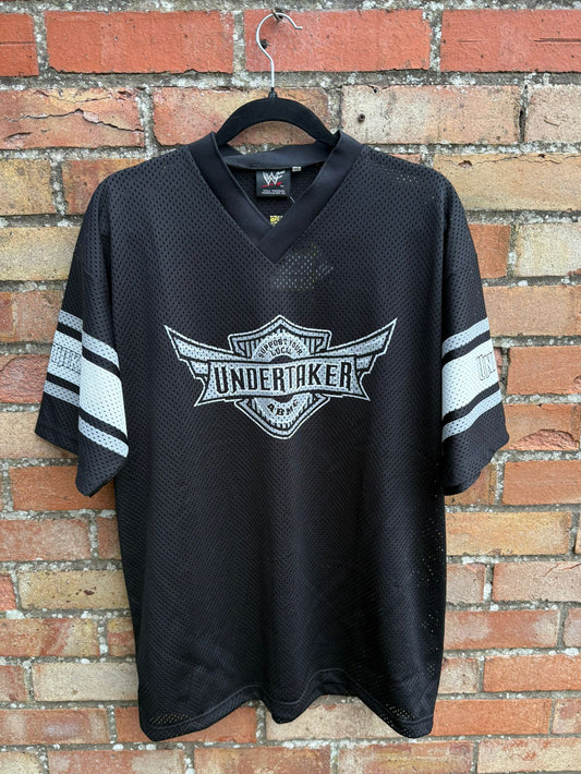 Black sports jersey with Undertaker logo on a hanger against a brick wall.