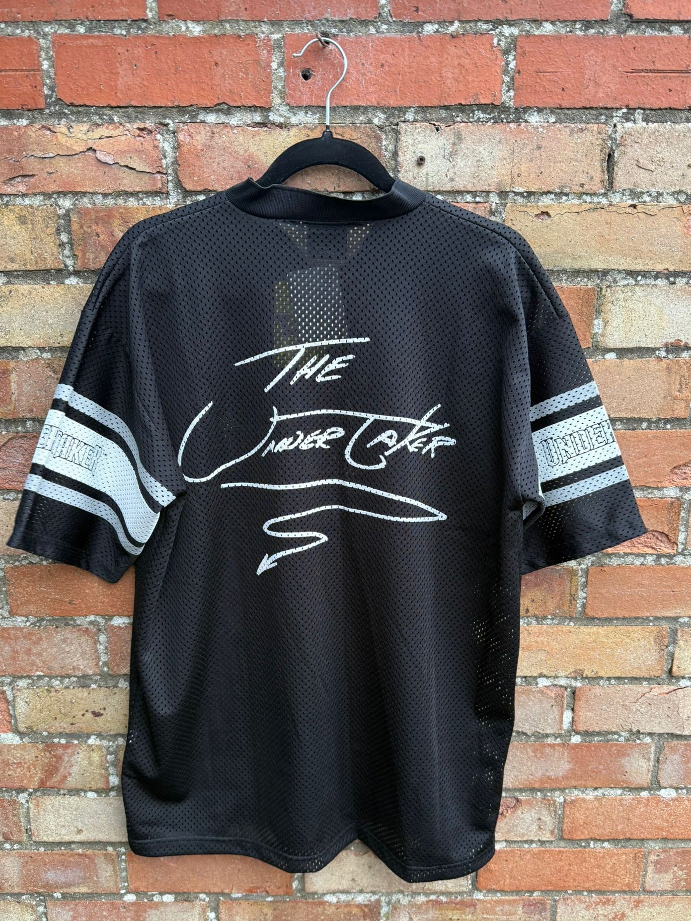 Back view of a black sports jersey with 'The Undertaker' text on a brick wall background