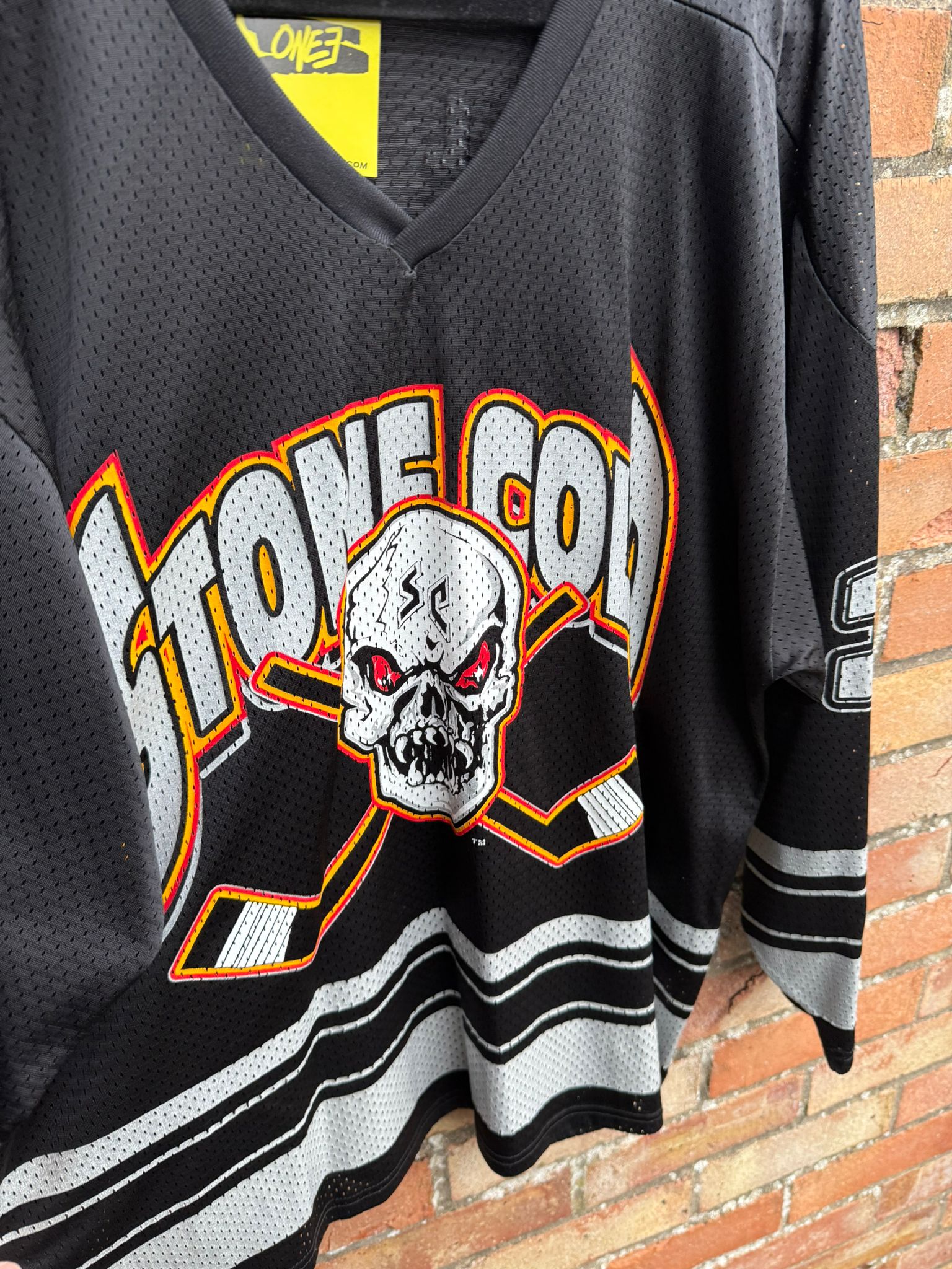 Black sports jersey with a skull with fangs design and 'Stone Cold' text against a brick wall.