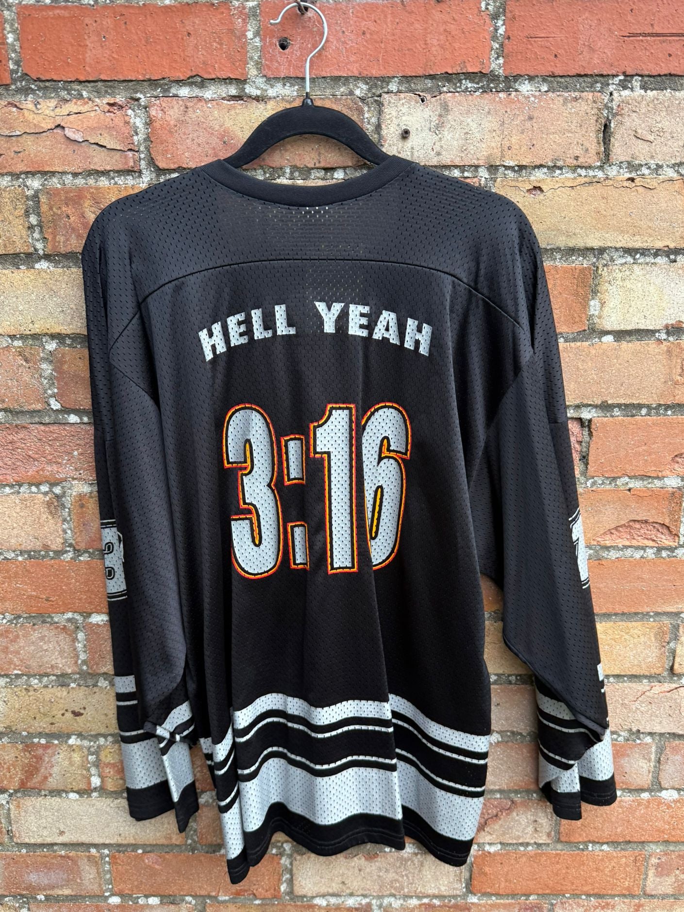 Back view of black sports jersey with 'HELL YEAH 3:16' text on a brick wall background