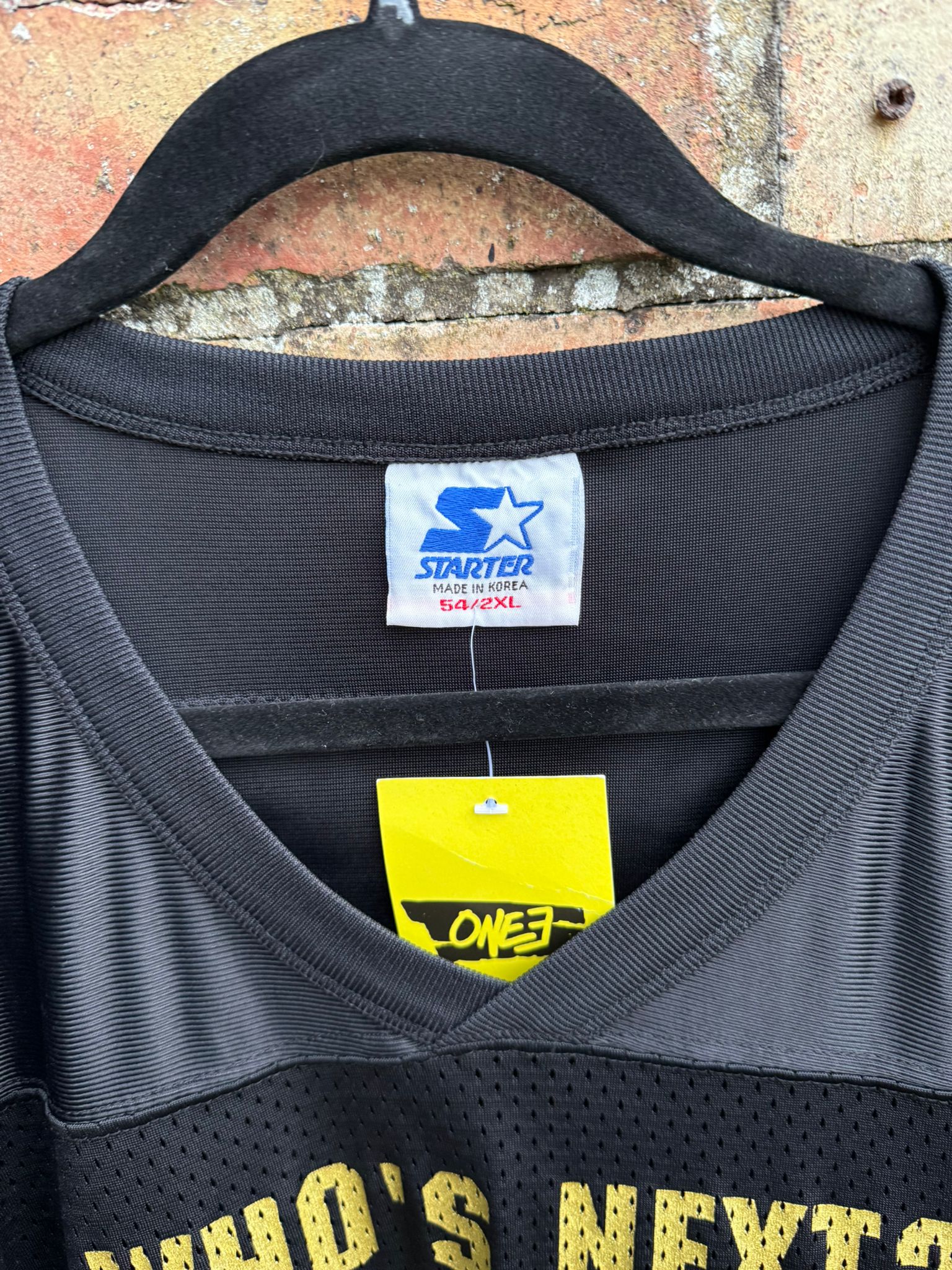Black sports jersey on a Starter tag with Who's next? in yellow text on a hanger against a brick wall.