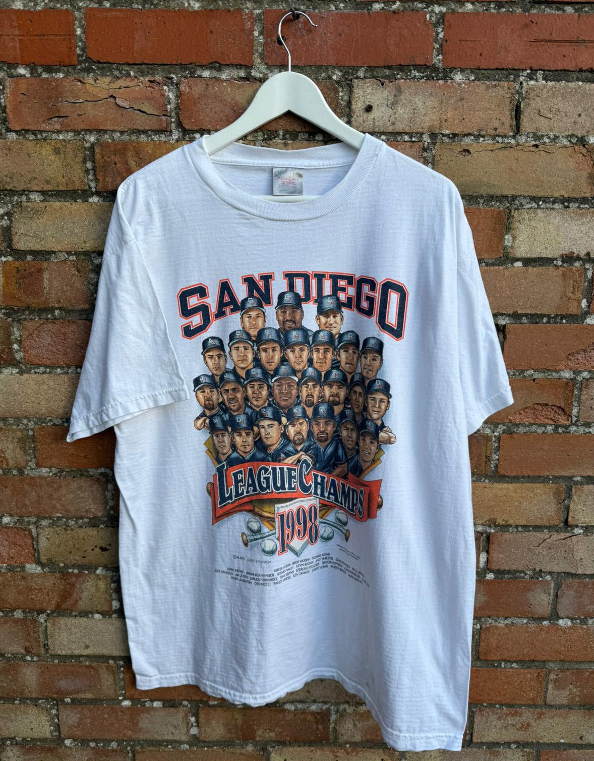 White t-shirt with San Diego Padres League Champs 1998 graphic on a hanger against a brick wall.