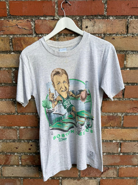 Gray t-shirt with a Boston Celtics graphic design on a hanger against a brick wall